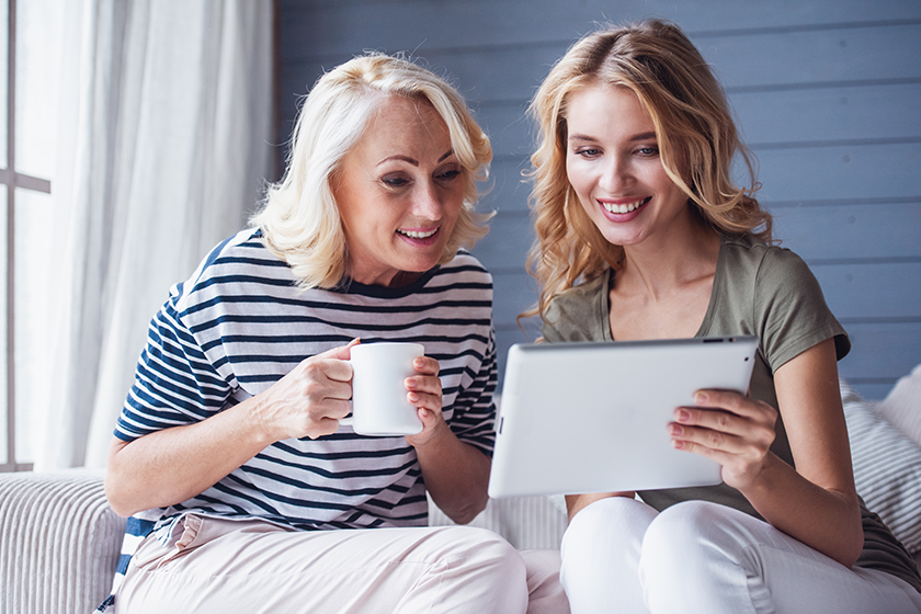 Beautiful senior mom and her adult daughter are using a digital tablet and smiling while sitting on couch at home 